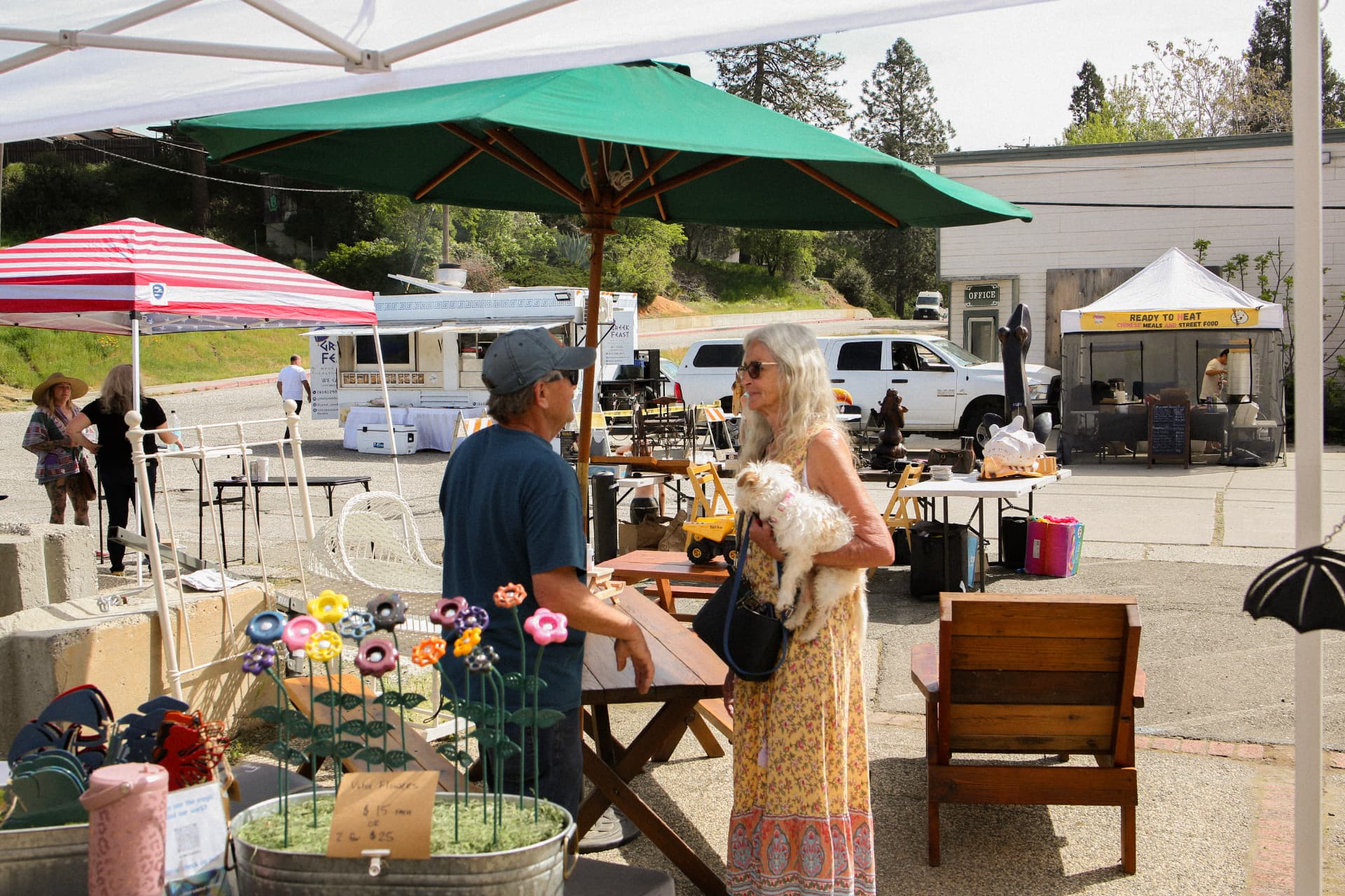 Food trucks and vendor booths in the lot at The Colfax Bazaar, with shoppers under a green umbrella.