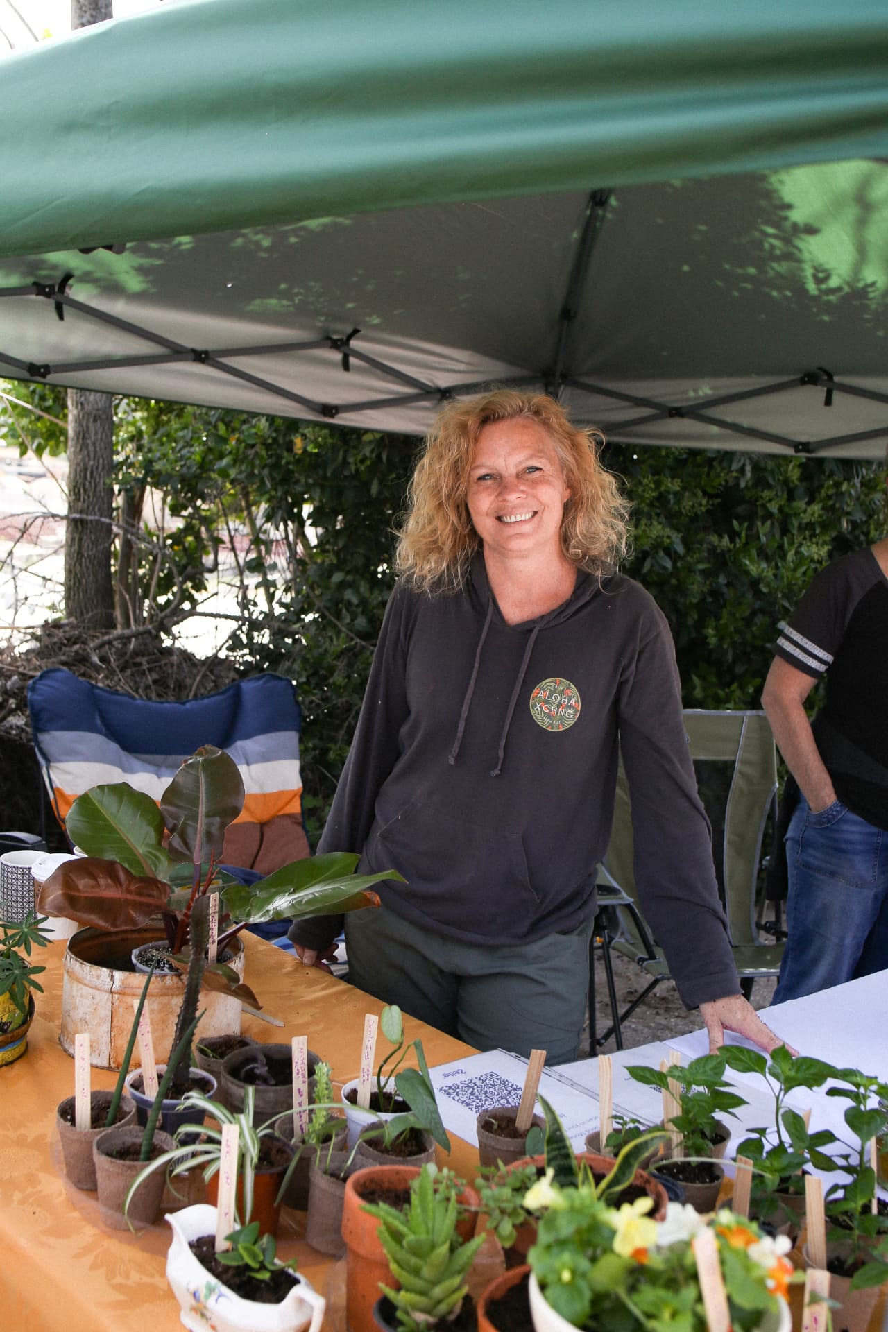 A smiling plant vendor behind a table of potted starts and houseplants under her canopy.